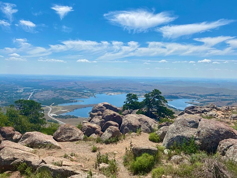 View of plains landscape with a shining lake from a mountain overlook; includes boulders in the foreground under a blue sky with bright white clouds (shot taken from Mount Scott, Wichita Mountains Wildlife Refuge, Oklahoma)