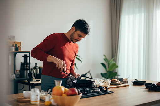 Handsome cheerful man preparing pancakes in the kitchen. He is standing wearing a red sweater.