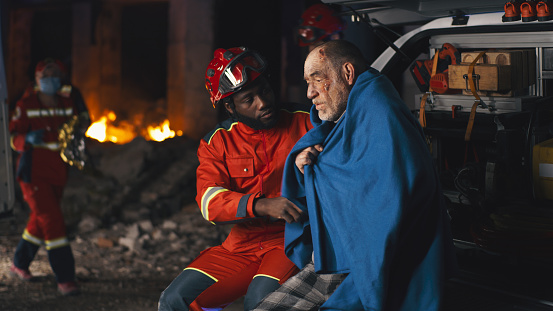 African American bearded man in paramedic uniform asking questions to senior man wrapped in blanket after accident at night