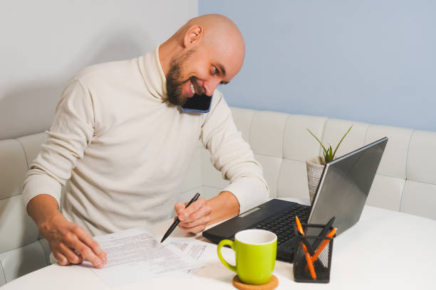 istock Smiling bald man with beard in white turtleneck working at home on notebook and speaking phone, side view 1356644495