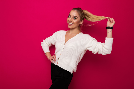 Photo of a beautiful pretty amazing positive smiling happy young blonde woman with makeup and ponytail in a stylish white knitted sweater and black pants isolated on a pink background with copy space for text.