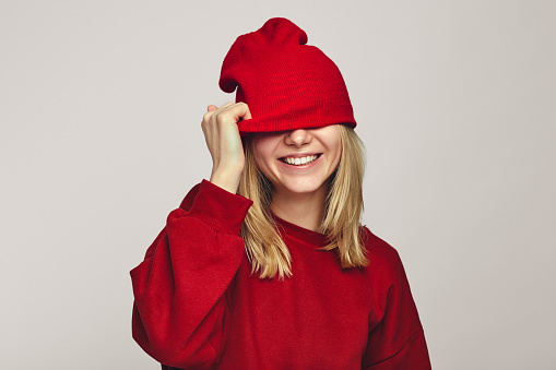 Happy faceless woman hides eyes with red hat wears stylish sweatshirt, feels glad isolated over white background. Season and clothes concept