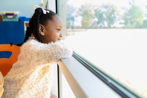 An adorable little girl daydreams while looking through a window in her kindergarten classroom.