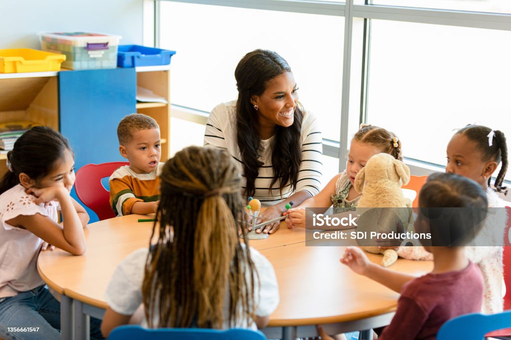 Smiling teacher teaches children about the solar system A cheerful mid adult preschool or kindergarten teacher smiles as she teaches a group of children about the solar system. A little girl is playing with a solar system model. Teacher Stock Photo