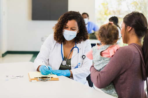 A concerned young mother talks with a female pediatrician during an appointment at a free clinic. The pediatrician is taking notes as the young mom talks about her daughter's symptoms.