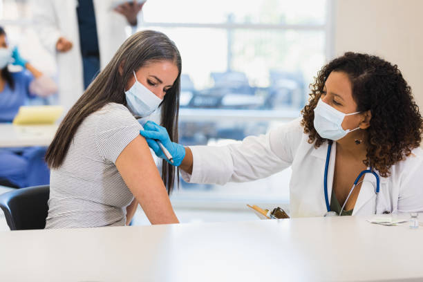 Woman prepares to receives Covid booster dose A mid adult female doctor prepares to administer a COVID-19 vaccine or booster dose to a mid adult female patient. The doctor and patient are wearing protective face masks. booster dose photos stock pictures, royalty-free photos & images