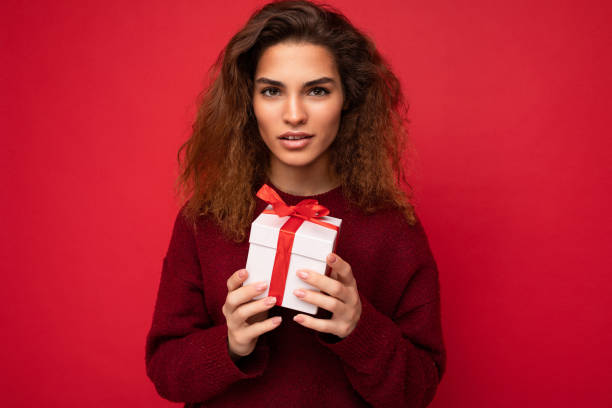 istock Charming self-confident young brunette curly woman standing isolated over red background wall wearing dark red sweater holding gift box looking at camera 1356669755
