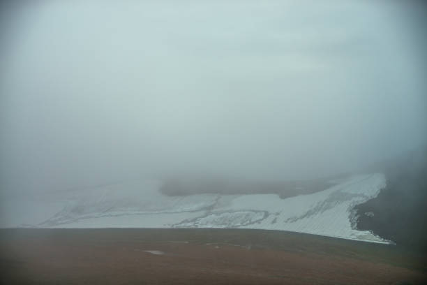 Atmospheric minimalist mountain landscape with small glacier on rocky hill slope inside low cloud. Mountainside with small glacier in dense fog. High mountain valley in low cloud. Poor visibility.