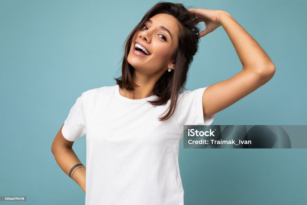 Photo of positive happy smiling young fascinating beautiful attractive nice brunette woman with sincere emotions wearing casual white t-shirt for mockup isolated over blue background with copy space for text Photo of positive happy smiling young fascinating beautiful attractive nice brunette woman with sincere emotions wearing casual white t-shirt for mockup isolated over blue background with copy space for text. T-Shirt Stock Photo