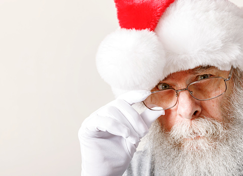 Santa Claus is peering intently over his eyeglasses which he is holding with his white-gloved hand.