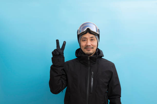Handsome Asian man with snowboard goggles and black anorak coat pointing finger on blue isolated background. Winter sport. Handsome asian man with snowboard goggles and black anorak coat pointing finger on blue isolated background. japanese snowboarder stock pictures, royalty-free photos & images