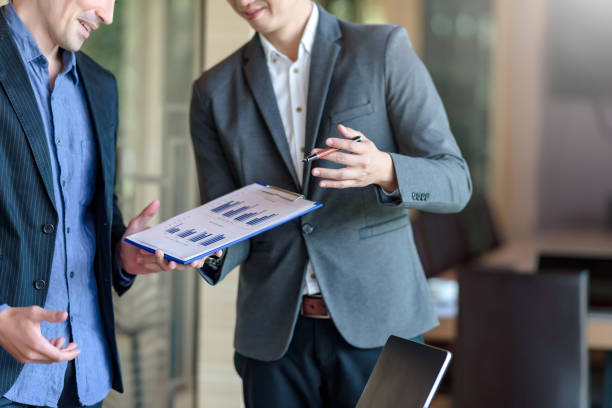 Two businessmen standing discussing with financial data and report graph in the office. Two businessmen standing discussing with financial data and report graph in the office. shareholder stock pictures, royalty-free photos & images