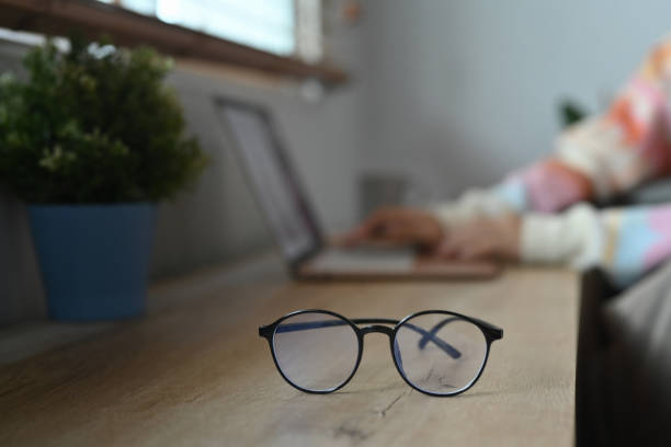istock Eyeglasses on wooden table and young woman using laptop in background. 1356711264