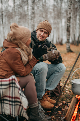 A loving couple in the fall on a camping, sitting on chairs near the fire, drinking tea. Blonde girl in a beige hat, brown jacket, brown leggings, gray boots. A man in a brown hat, khaki jacket, jeans and brown boots.