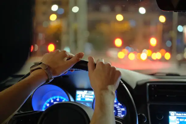 Photo of Close up of driver hands holding steering wheel driving car with blurred city street lights on background at night