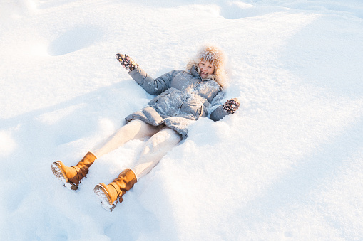 Attractive smiling mid adult Caucasian woman wearing gray down jacket and yellow boots lying in snow making snow angel.