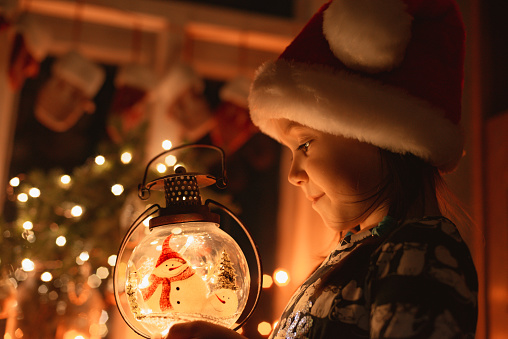 little girl looks at a Christmas lantern illuminating the face in Santa's hat at night in the room, child makes a wish for the New Year and dreams of a gift