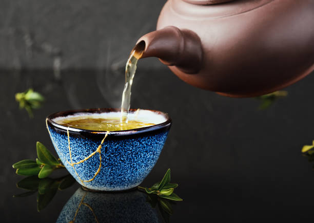 Hot green tea is poured from a ceramic teapot into a bowl. Selective focus on the blue cup. Steam will rise above the mug Reclaimed ceramic blue cup, second life of things, recycling or kintsugi stock photo