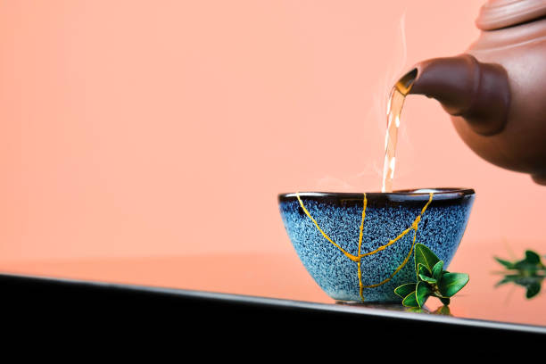Hot green tea is poured from a ceramic teapot into a bowl. Selective focus on the blue cup. Steam will rise above the mug Reclaimed ceramic blue cup, second life of things, recycling or kintsugi stock photo