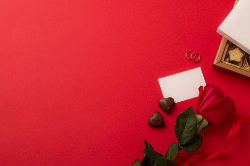 Top view of two wedding rings surrounded by a red rose, a greeting card, two heart-shaped chocolates a red ribbon and a a chocolate box on a red background. All the objects are at the right side of the image so there is a useful copy space on the greeting card and at the left side on the red background