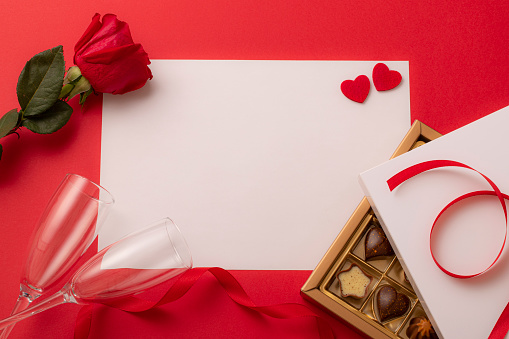 Top view of a white paper framed by a red background with two hearts shape, two champagne flute, a chocolate box, a red rose and a red ribbon. There is a useful copy space at the image on the white background.