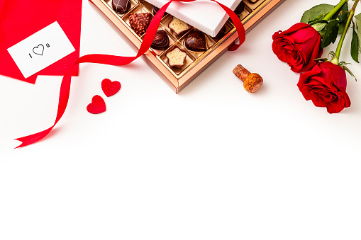 Top view of a opened chocolate box with a red ribbon surrounded by two red roses, a champagne cork, two hearts shape and a greeting card. All the objects are at the top of the image leaving a useful copy space at the lower side on a white background.