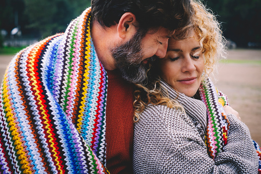 Romantic adult couple stay together with feeling and love covered by a woolen colorful blanket at the outdoor park. Concept of happy relationship lifestyle people. Winter leisure season outside