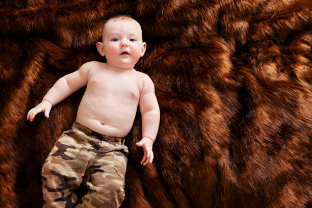 un drôle de petit garçon heureux est allongé sur un tapis de fourrure brune dans un uniforme militaire. enfant souriant au ventre nu en vêtements kaki, vue de dessus - fake fur white happiness beginnings photos et images de collection