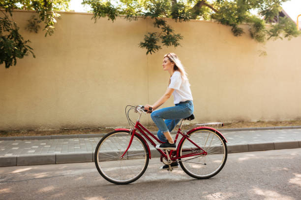 幸せに自転車に乗ってブロンドの髪を持つ美しい女の子。街の通りに沿って赤い自転車に乗っている間楽しんで頭の上にジーンズとサングラスの若い女性 - women t shirt smiling isolated ストックフォトと画像