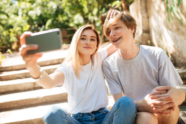 若いカップルは公園の階段に座って、一緒に面白い自分撮りを作ります。携帯電話の正面カメラで写真を撮りながら、カメラを見てブロンドの髪を持つ笑顔の男の子と美しい女の子 - women t shirt smiling isolated ストックフォトと画像