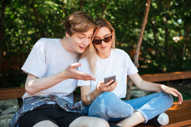 istock Joyful couple sitting on bench in park and emotionally discussion something while using cellphone. Close up photo of young smiling man and nice lady with blond hair spending time together 1356755227