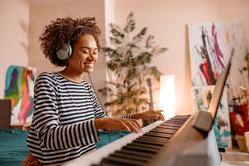 Smiling multiracial female musician in headphones playing melody on synth electronic musical instrument