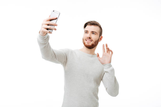 istock Cheerful handsome man taking selfie and waving hello to the front camera over white background 1356758472