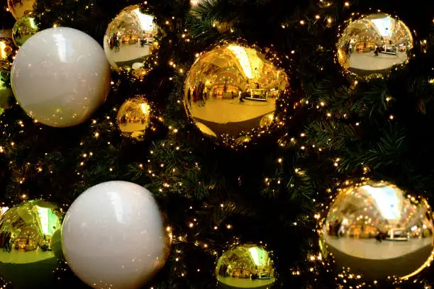 decorative shiny large reflective gold and white sphere shape Christmas ornaments. artificial green plastic tree with fine needles. strong reflections. yellow lights. seasonal. interior space. closeup view.