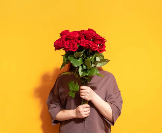 Photo of In female hands a bouquet of fresh red roses. The faceless trend. Yellow background. Selective focus.