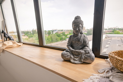 Buddha Figurine in Meditation Pose on a Wooden Windowsill Next to a Large Window. Close-up . High quality photo