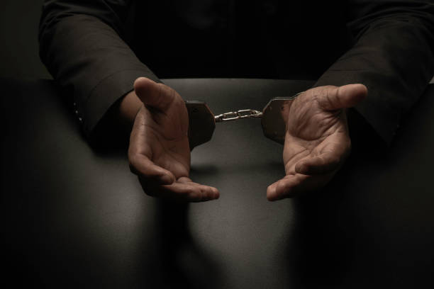 istock A businessman in a black suit, handcuffed on a table and a black table, is making gestures by raising his hand explaining to the police being released. Corruption, bribery, illegal transactions. 1356771983