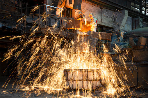Male foundry worker creating sparks pouring molten metal in the container.