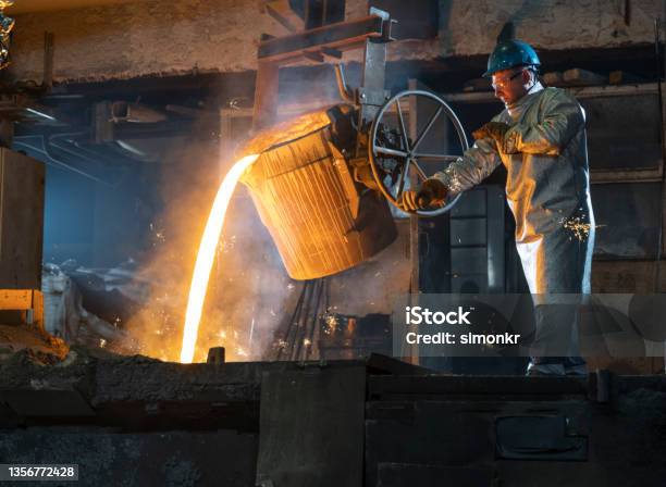 Foundry Worker In Protective Coat Pouring Molten Metal From The Ladle Stock Photo - Download Image Now