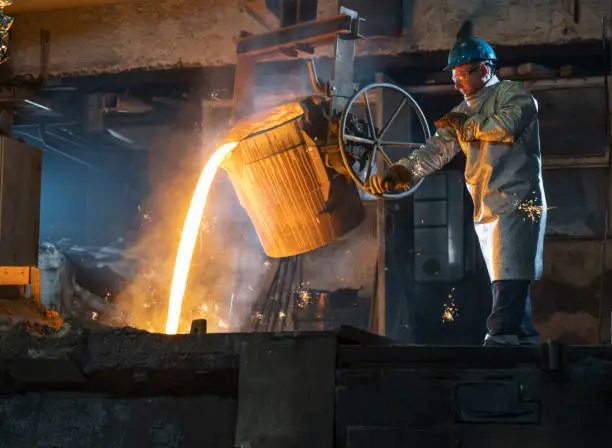 Mature male worker wearing protective coat while pouring molten metal at the foundry.
