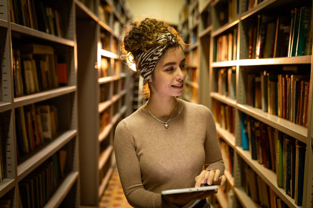 istock Young female student at library trying to find book while using tablet 1356776997