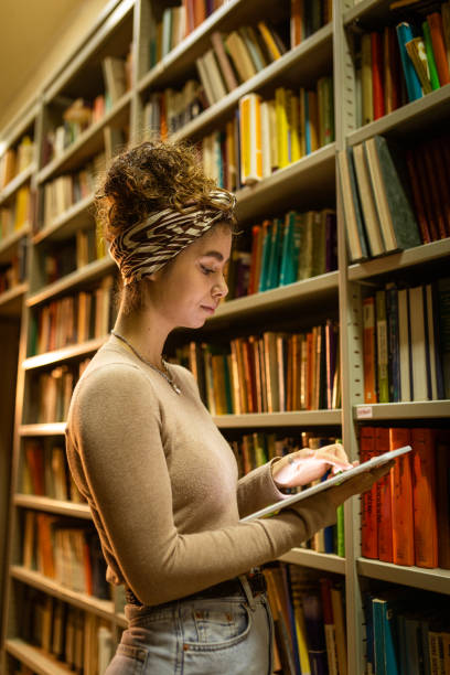 istock Young female student at library trying to find book while using tablet 1356777046