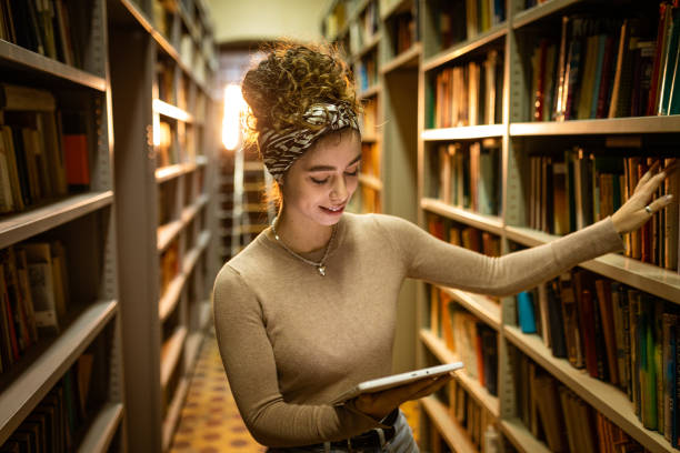 istock Young librarian picking book from bookshelf and holding tablet 1356777975