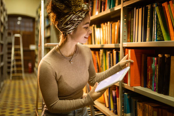 istock Young librarian picking book from bookshelf and holding tablet 1356777989