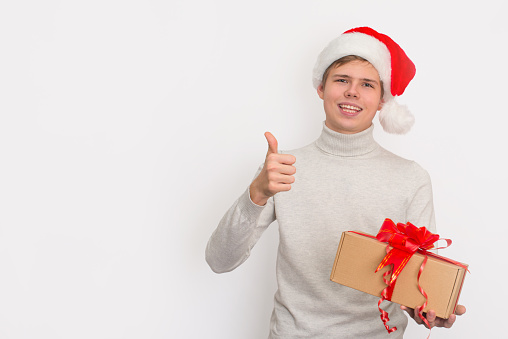 Teenager boy in Santa hat holding Christmas present  and showing thumb up gesture on white background with copy-space