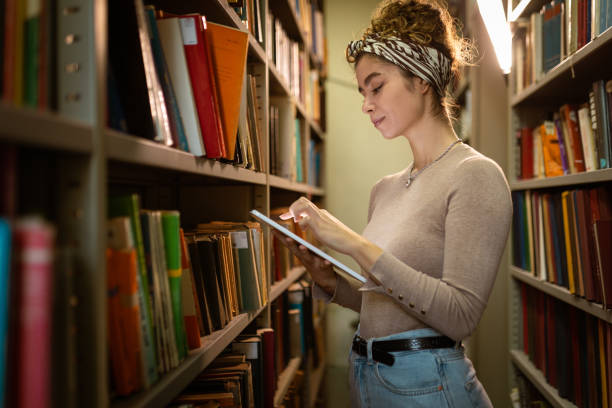 istock Young student holding tablet and searching book at library 1356778089
