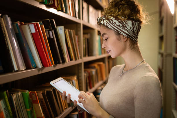 istock Young student holding tablet and searching book at library 1356778123
