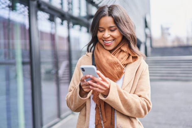 mensajería de mujer con abrigo en un teléfono inteligente de pie afuera en la ciudad - usar el teléfono fotografías e imágenes de stock