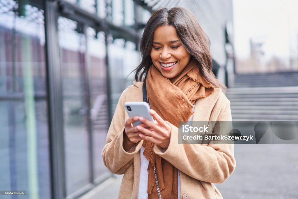 Woman in coat messaging on smartphone standing outside in the city Using Phone Stock Photo