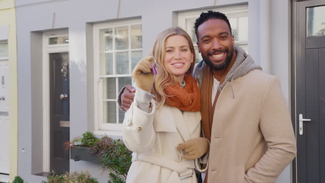Portrait of multi cultural couple outside house on moving day holding keys to new home in fall or winter - shot in slow motion